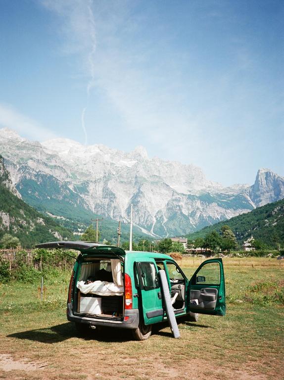 A green van with its doors open is parked in a grassy field with mountains in the background.