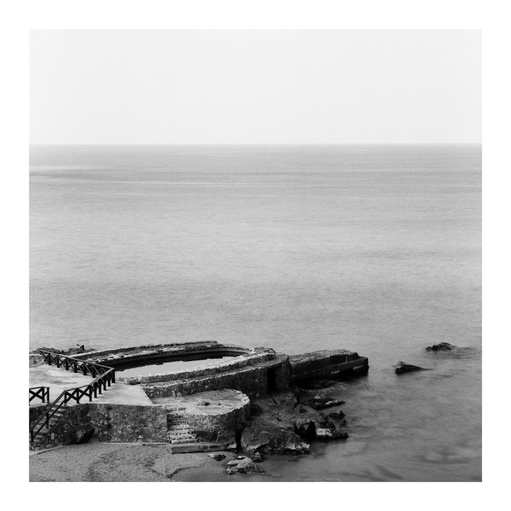 A concrete structure with a pool and wooden railing sits on a rocky shore next to the ocean.