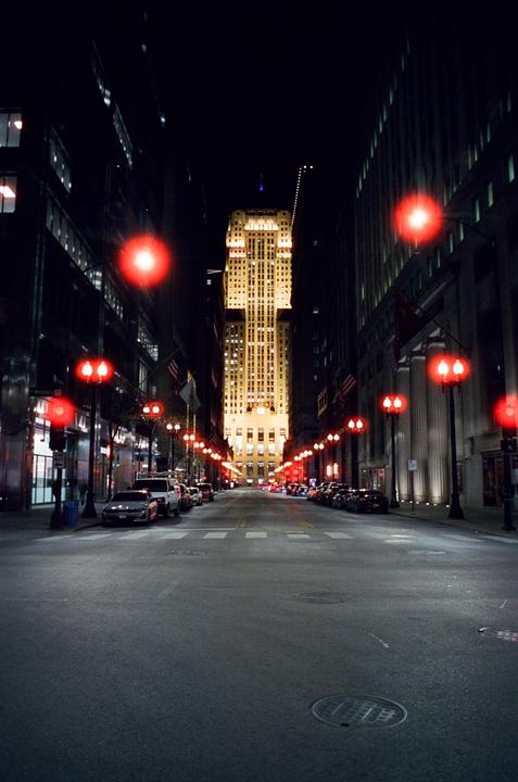 A street at night is lined with parked cars and red streetlights, leading to a brightly lit skyscraper in the distance.