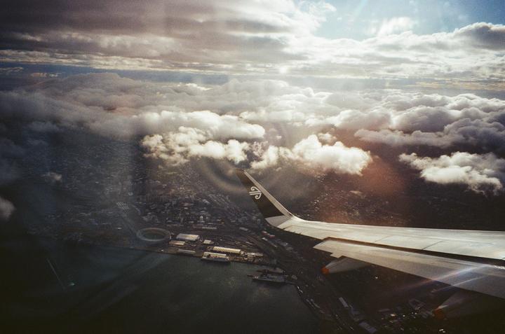 An airplane wing is visible over a city and harbor, with clouds and sun rays in the sky.