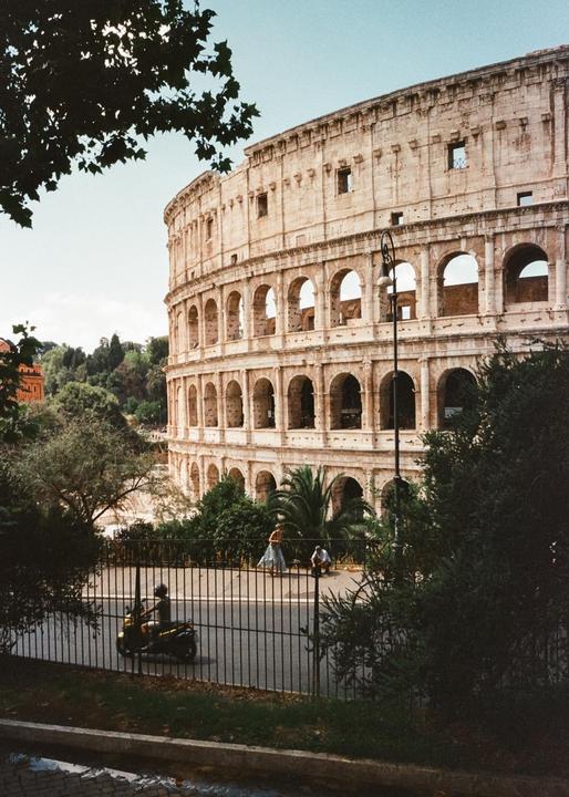 The Colosseum in Rome is visible behind a street with a scooter and people.