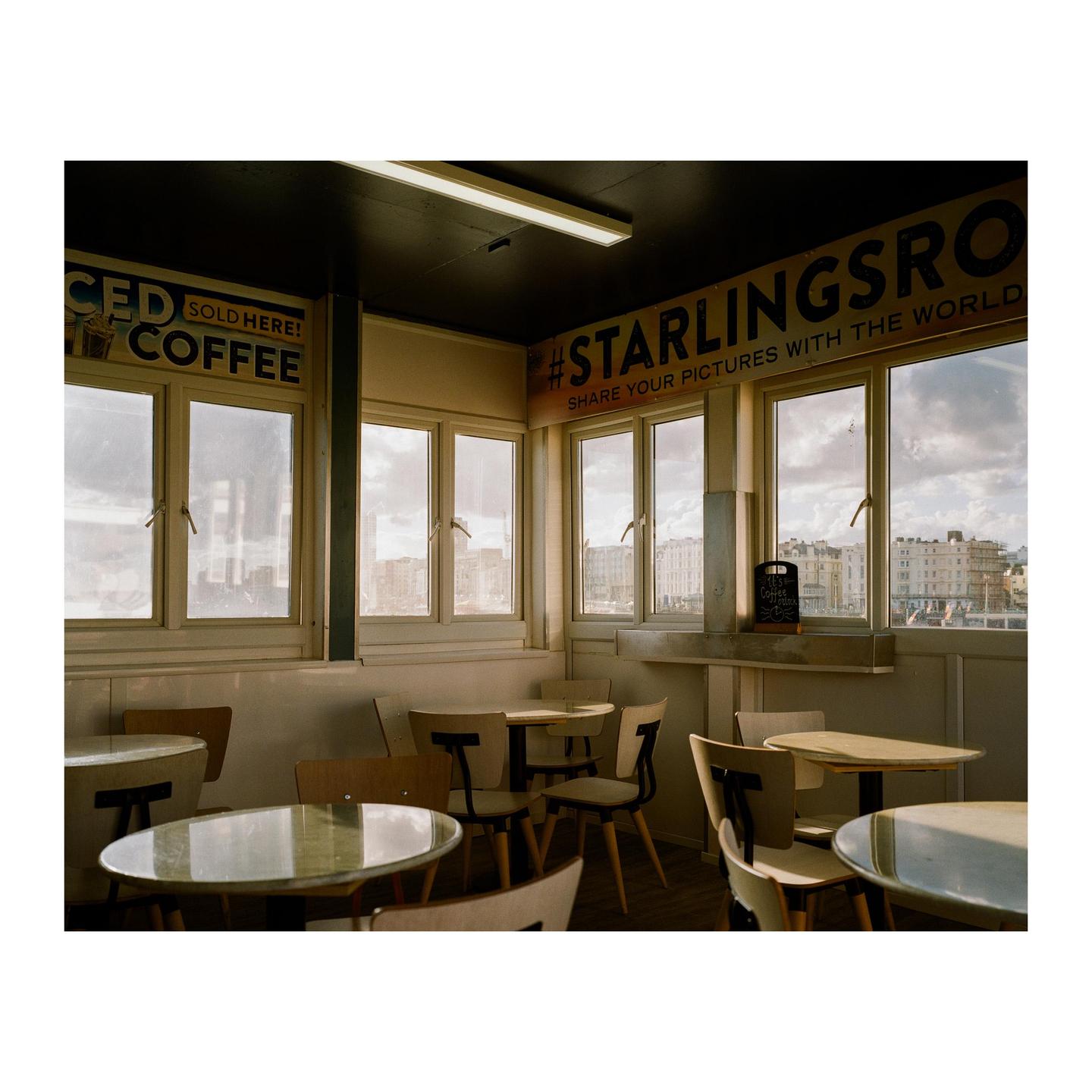 A cafe with many tables and chairs is illuminated by golden hour light coming through large windows overlooking a city.