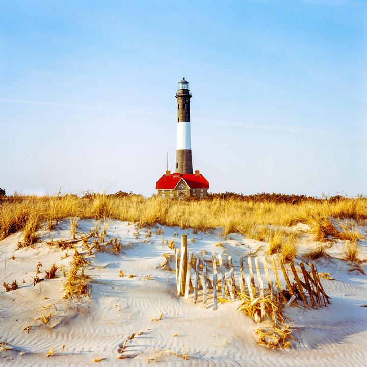 A lighthouse with a red roof stands behind sand dunes and dry grass under a clear sky.