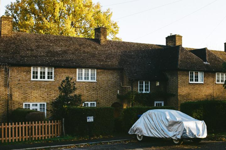 A car covered with a silver tarp is parked in front of a brick house with a hedge and a wooden fence.