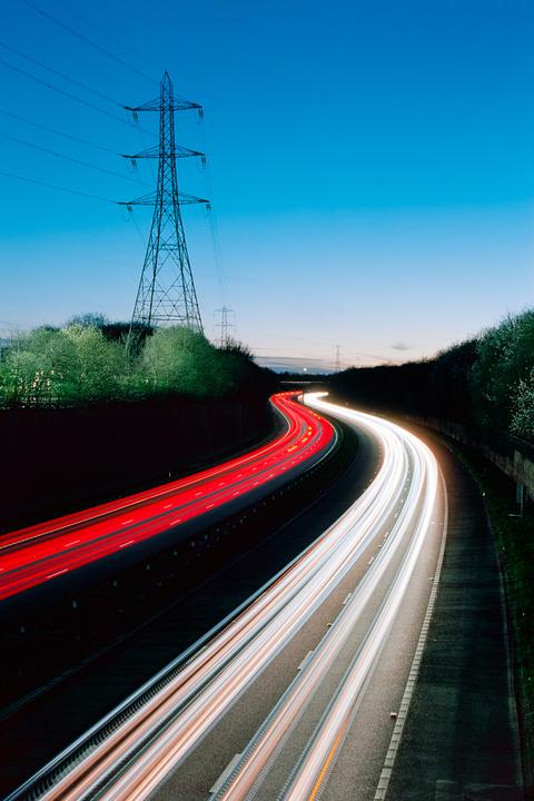 Light trails from vehicles on a highway at dusk with a large electricity pylon visible against the sky.