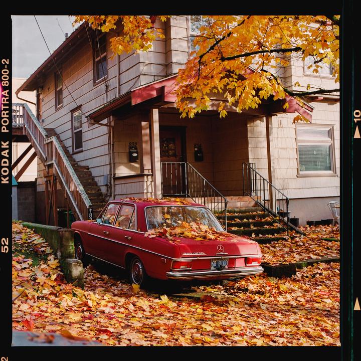 A red car is parked in front of a house, surrounded by autumn leaves.