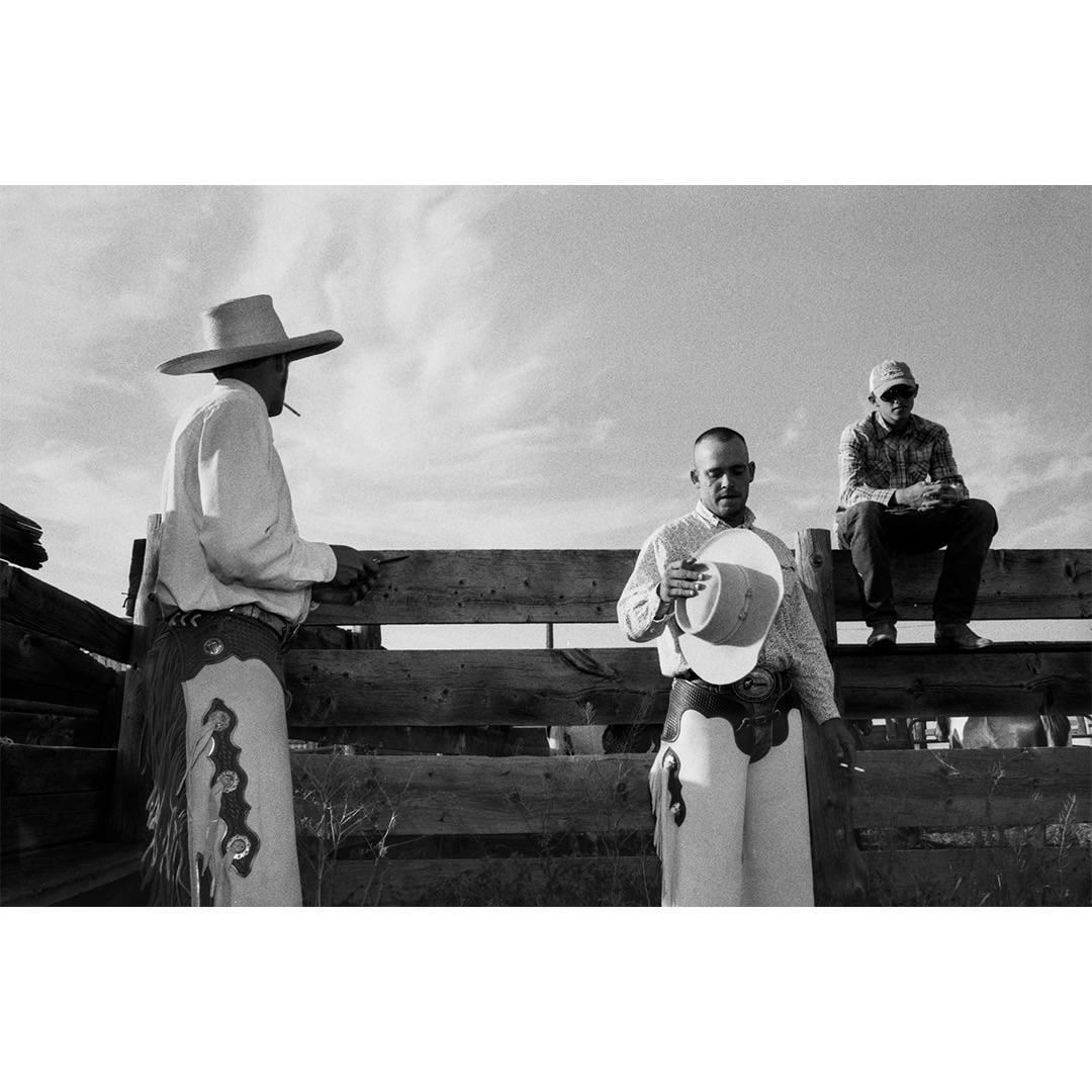 Three men in cowboy attire stand and sit by a wooden fence under a cloudy sky.