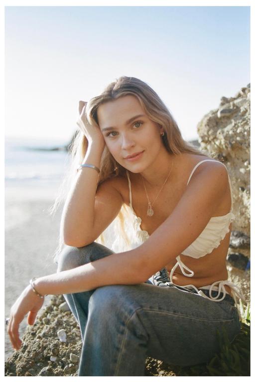 A woman with long blonde hair sits on a rocky beach, looking at the camera.