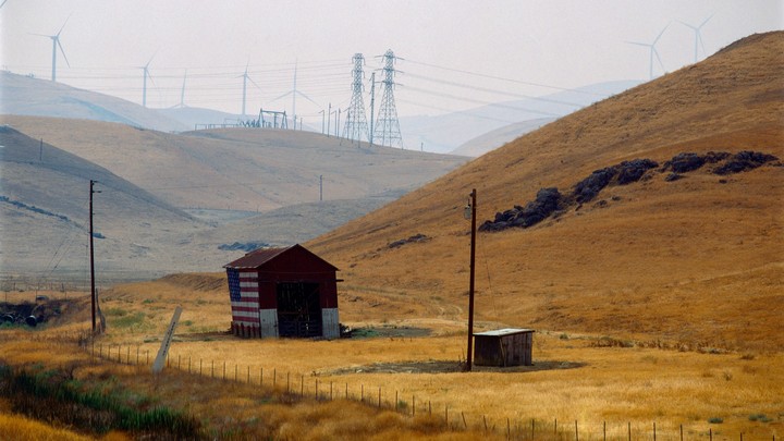 A small barn painted with an American flag sits in a dry, grassy valley with wind turbines and power lines in the distance.