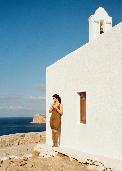 A woman in a brown dress stands next to a white building with a bell tower overlooking the sea and a rocky island.