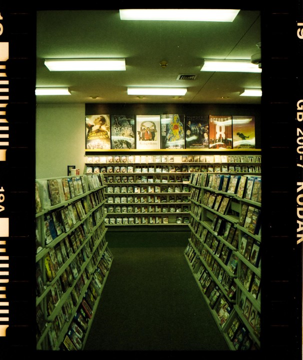 A narrow aisle in a video store is lined with shelves of movies and movie posters.
