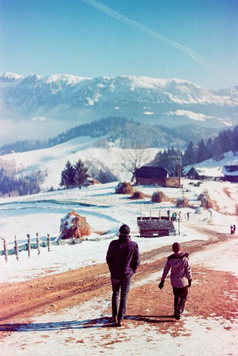 A man and a boy walk along a dirt road through a snow-covered mountain village with haystacks and houses.