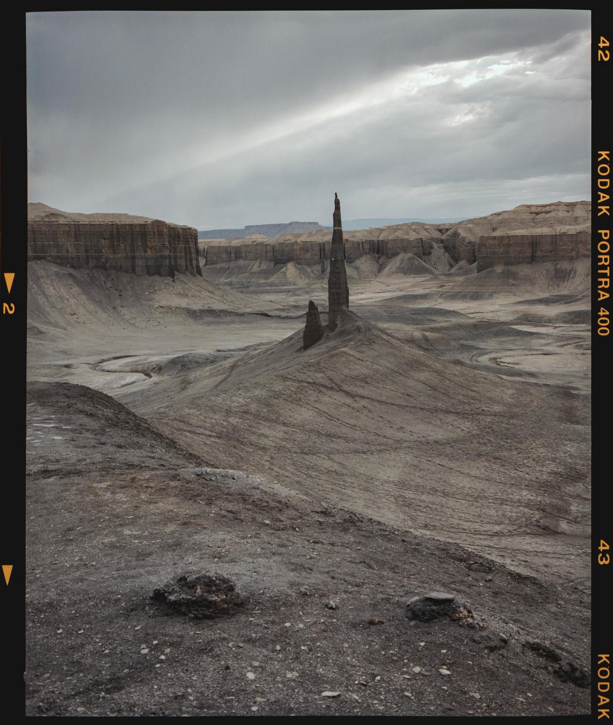 A tall, thin rock formation stands in a barren, rocky landscape under a cloudy sky.