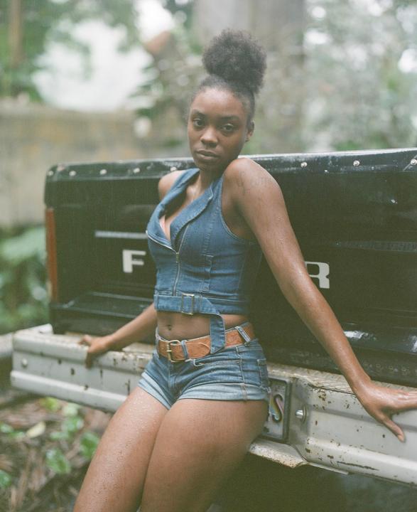A woman in denim shorts and a denim top is sitting on the tailgate of a truck.