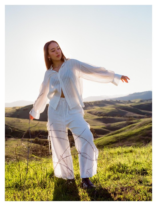 A woman in white clothing stands in a grassy field with rolling hills in the background.