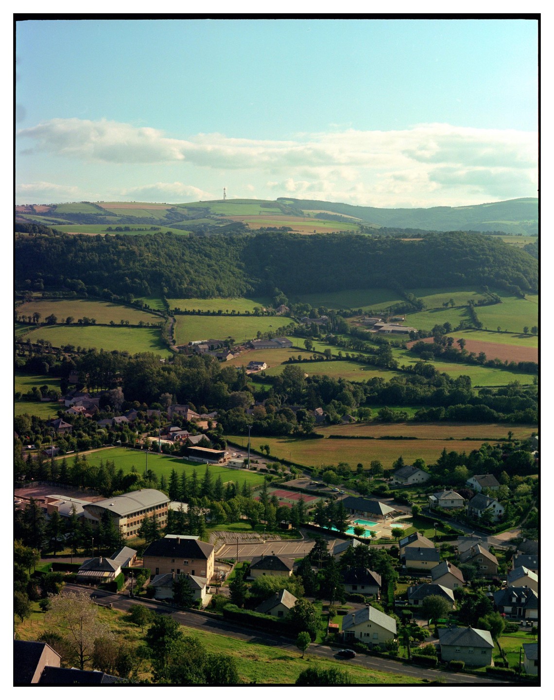 A small town with houses and buildings is nestled in a valley surrounded by green hills and fields under a partly cloudy sky.