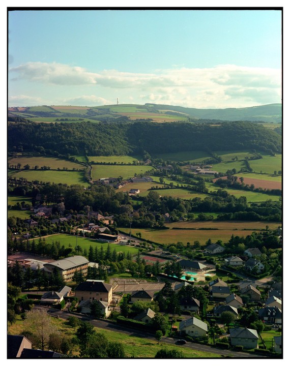 A small town with houses and buildings is nestled in a valley surrounded by green hills and fields under a partly cloudy sky.