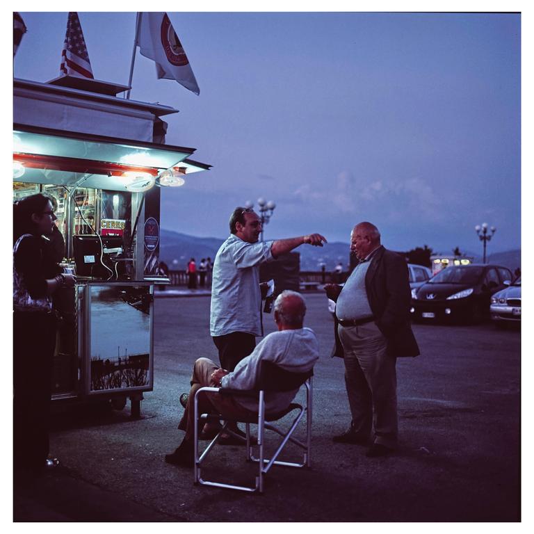 Three men are gathered around a food truck with an American flag and a white flag on top, with one man sitting in a folding chair and another pointing into the distance.