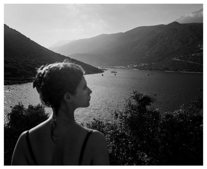 A woman looks out over a bay with mountains and boats in the distance.