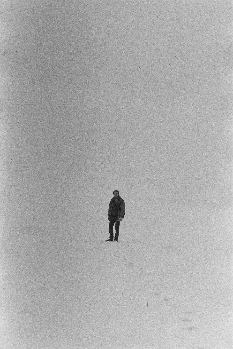 A man stands in a vast snowy landscape with footprints leading away from him.