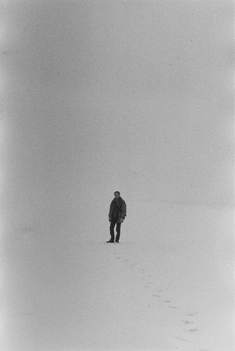 A man stands in a vast snowy landscape with footprints leading away from him.