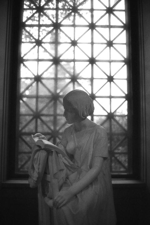 A statue of a woman reading a book is backlit by a window with a geometric grid.