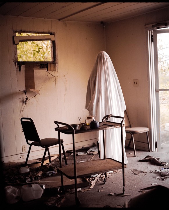 A person draped in a white sheet stands in a dilapidated room with a serving cart and chairs.