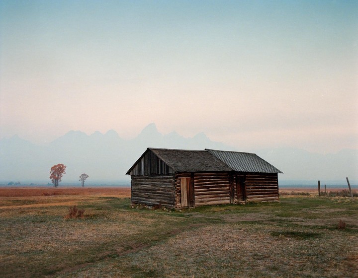 A wooden cabin sits in a field with mountains in the background under a hazy sky.