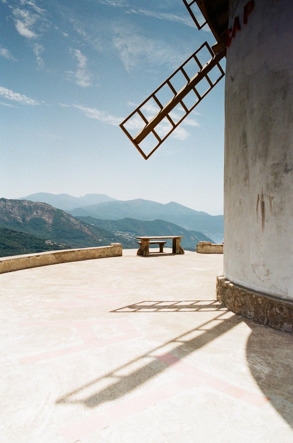 A windmill blade casts a shadow on a patio overlooking mountains and a body of water.
