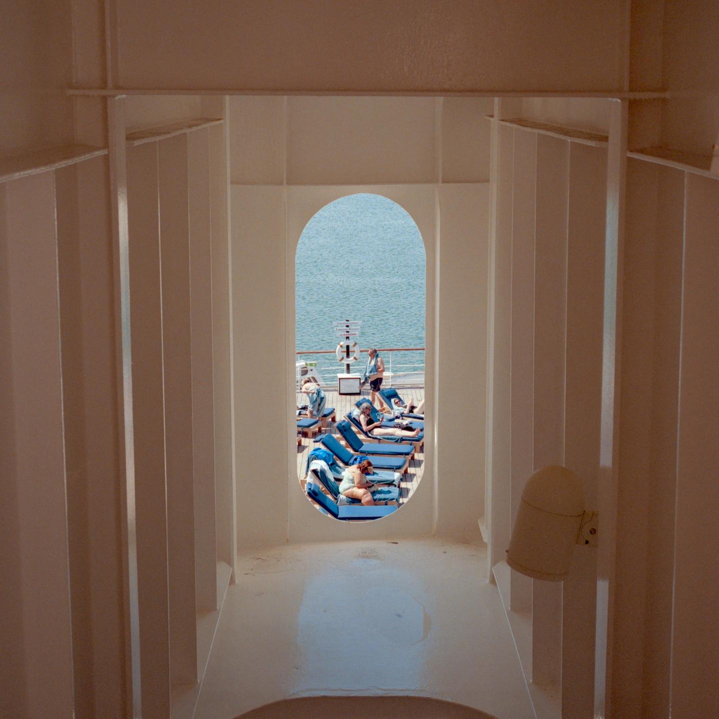 A view through an arched opening shows people relaxing on lounge chairs on a ship deck with the ocean in the background.