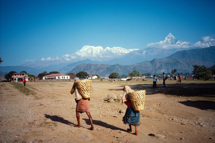 Two people with large woven baskets on their backs walk across a dirt field toward a village with snow-capped mountains in the distance.