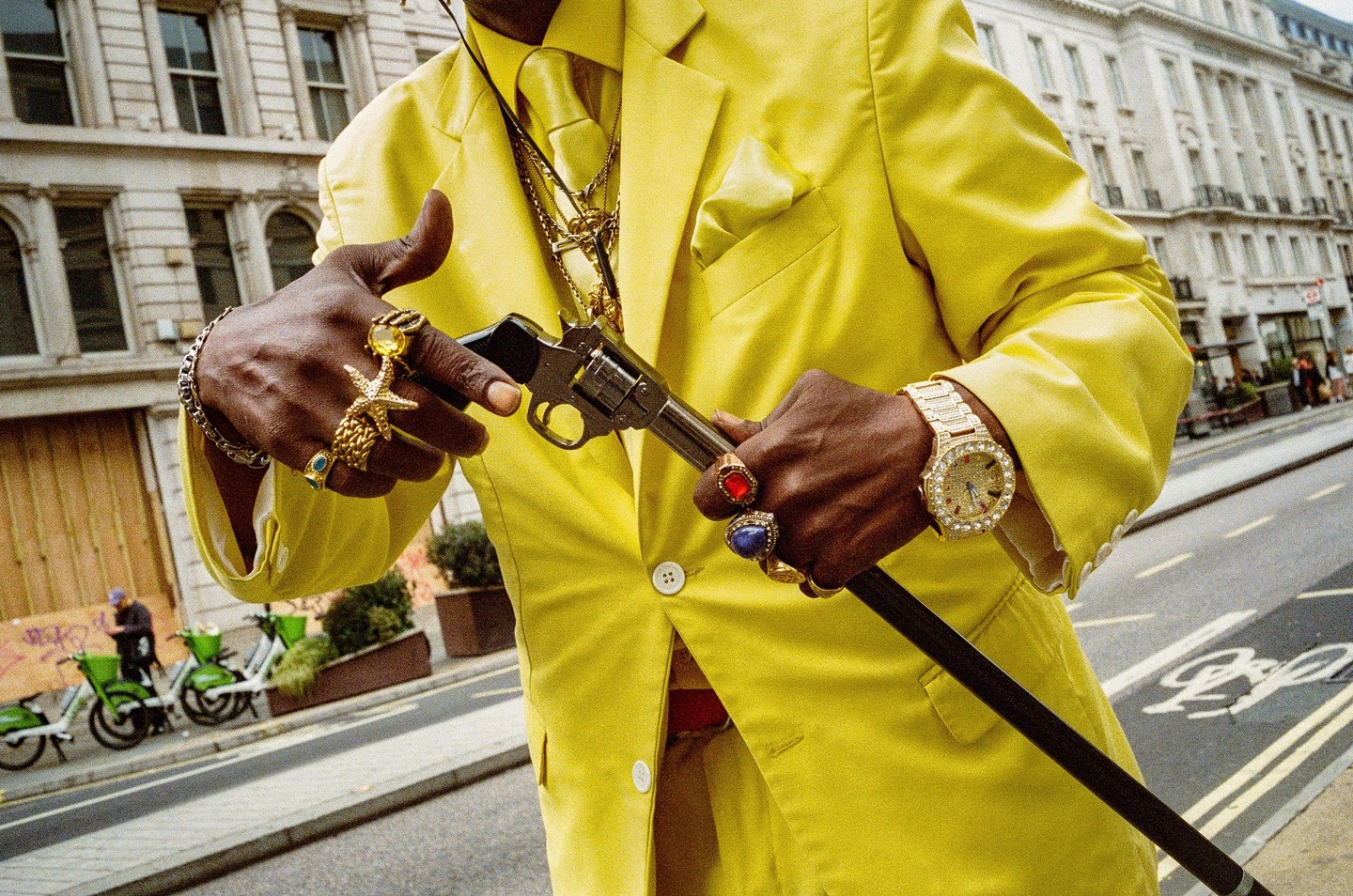 A man in a bright yellow suit holds a cane with a revolver handle, adorned with numerous rings and a gold watch.