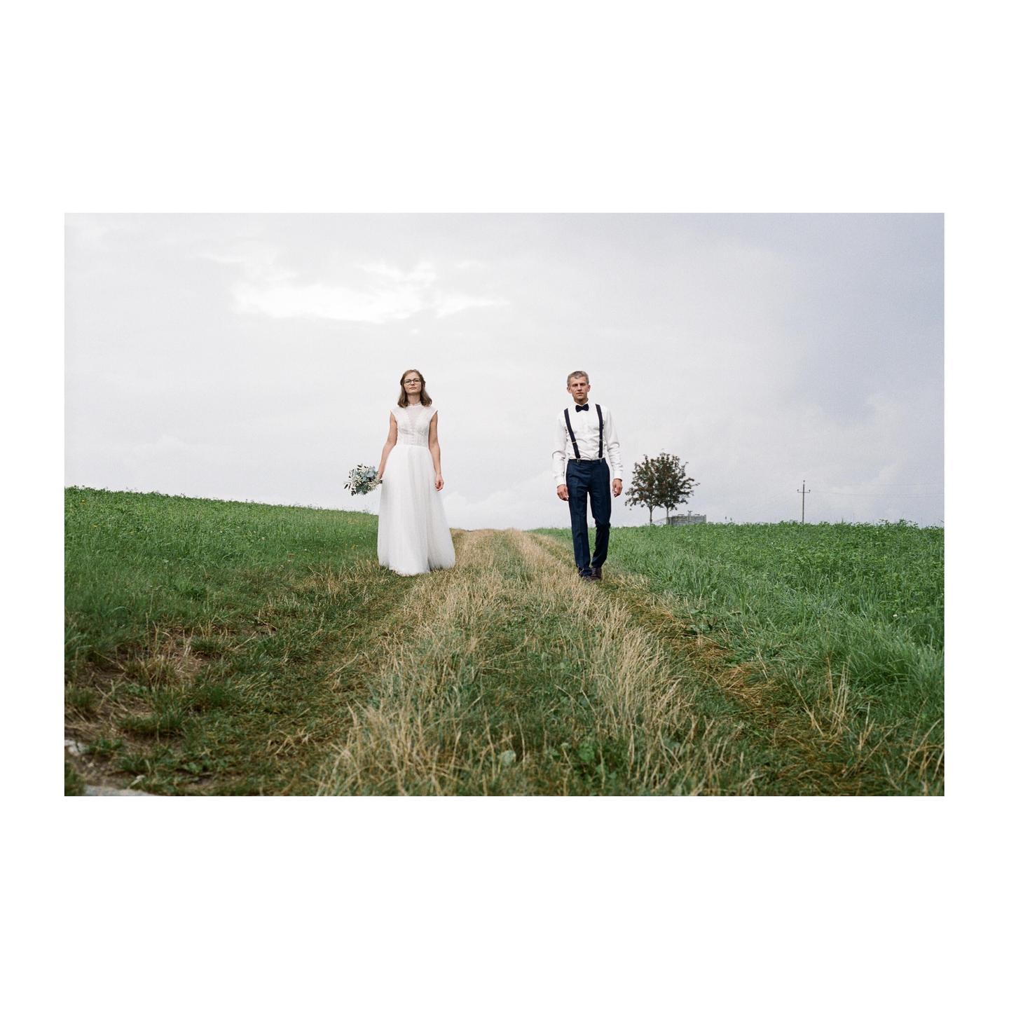 A couple walks on a dirt path through a grassy field under a cloudy sky.