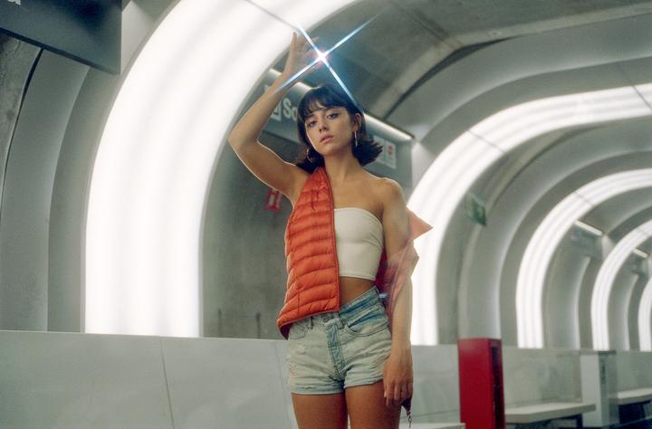A woman in a white tube top, denim shorts, and an orange vest stands in a subway station with her arm raised, catching a starburst flare from a light.