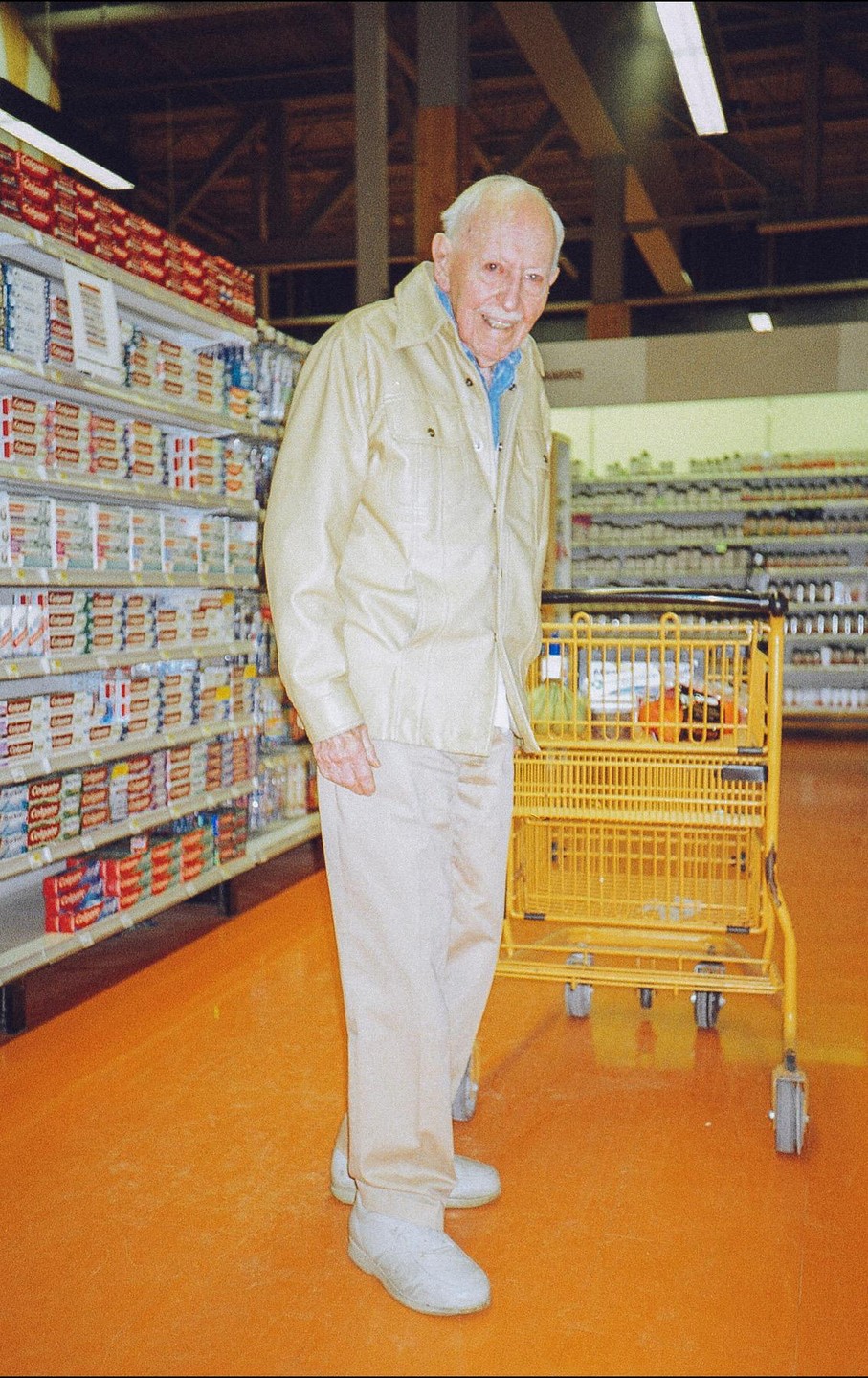A smiling elderly man stands next to a yellow shopping cart in a grocery store aisle.