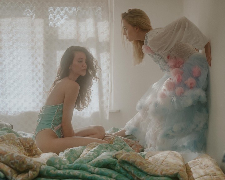 Two women are in a bedroom, one sitting on a bed and the other leaning against a wall.