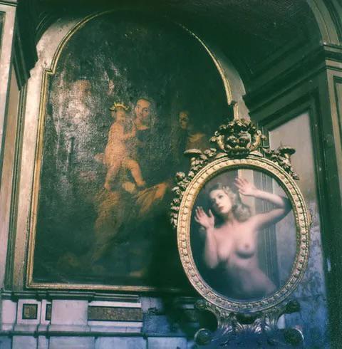A nude woman is reflected in an ornate mirror next to a painting of a child and two adults.