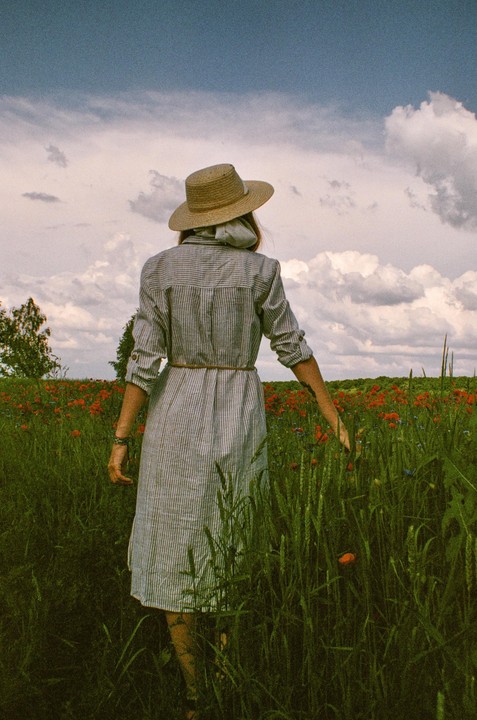 A woman in a striped dress and straw hat walks through a field of poppies under a cloudy sky.