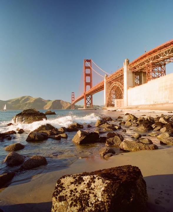 The Golden Gate Bridge is visible from a rocky beach with waves crashing on the shore.