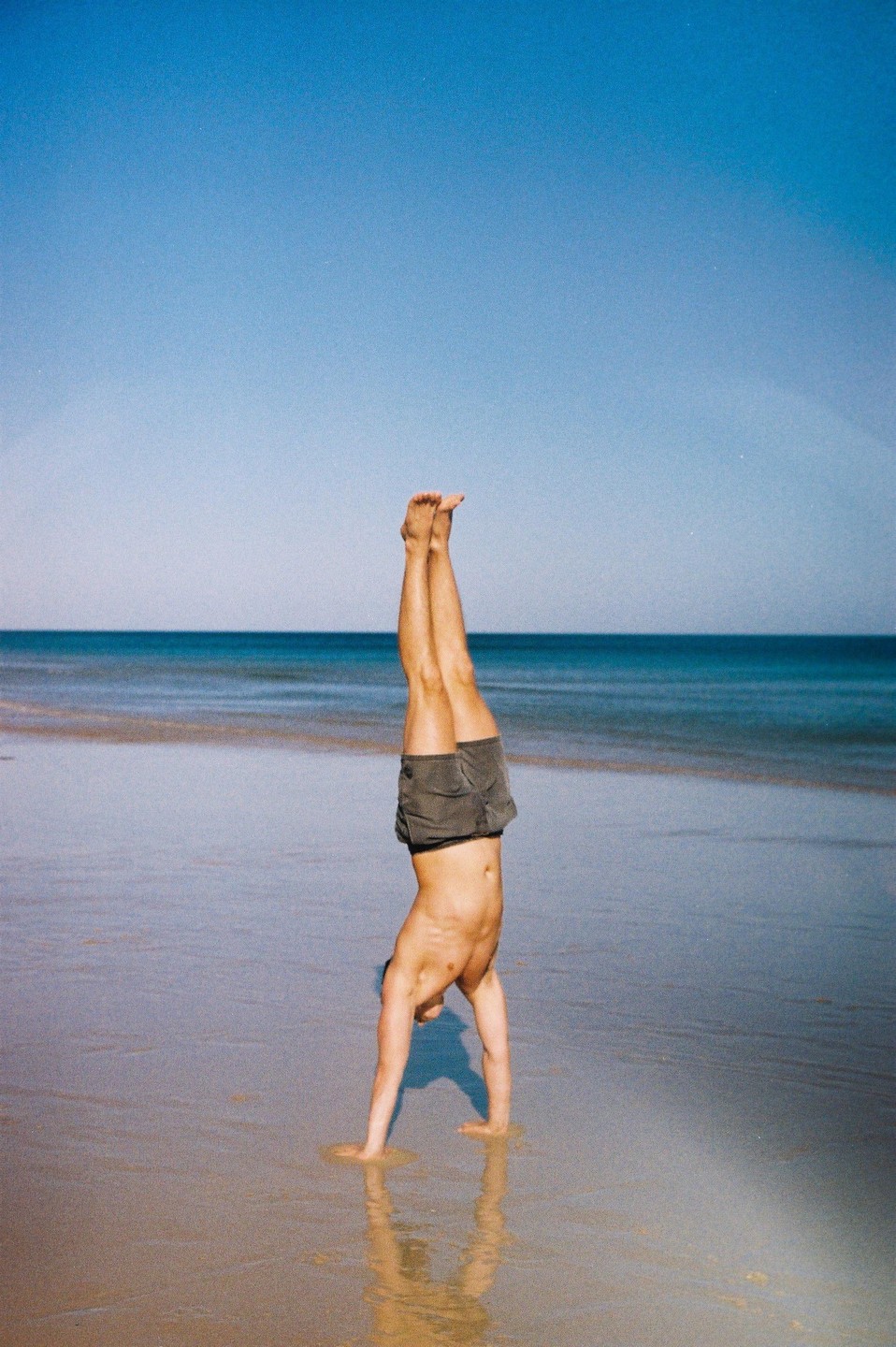 A man performs a handstand on a wet sandy beach with the ocean and clear sky in the background.