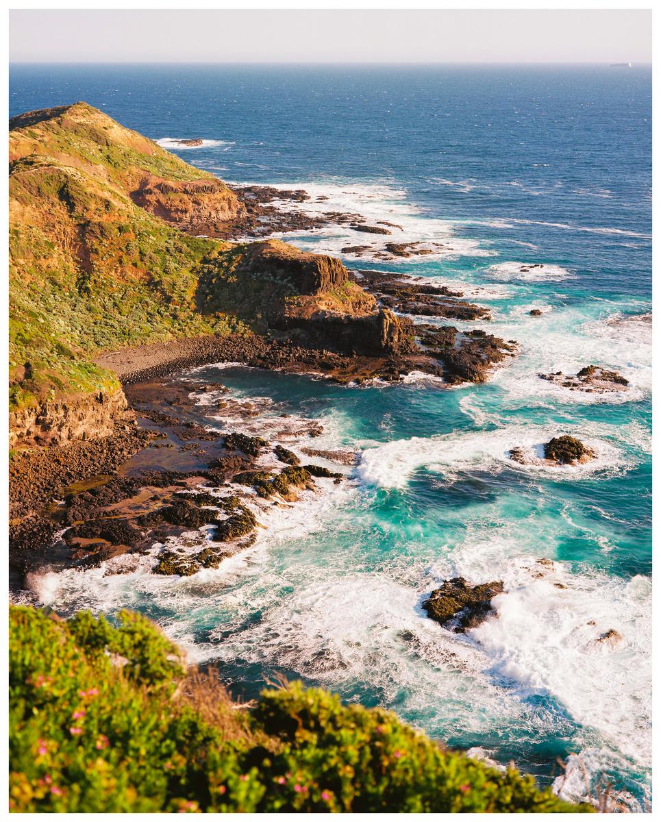 A high-angle landscape view shows a rocky coastline with green hills meeting a turquoise ocean with white waves.