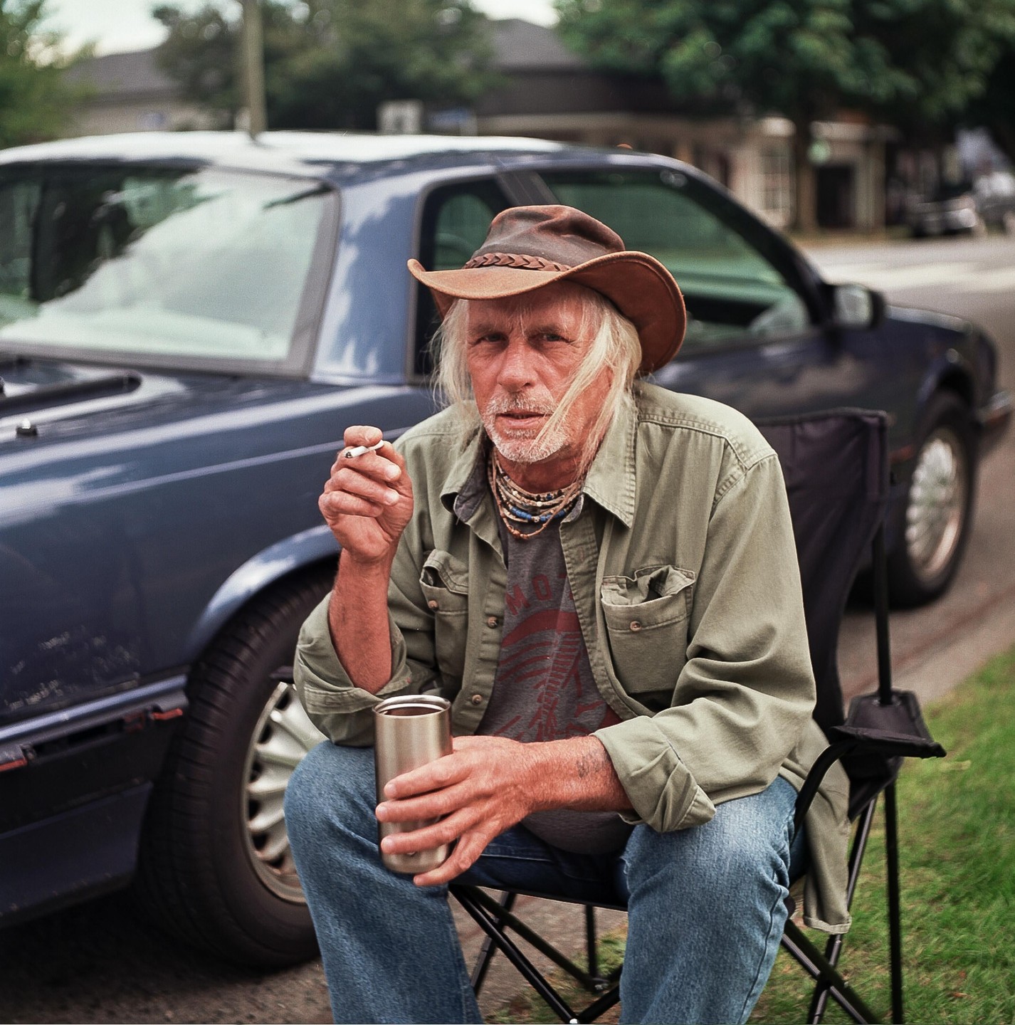 A man with long white hair and a cowboy hat sits in a folding chair on the side of a street, holding a cigarette and a metal cup.