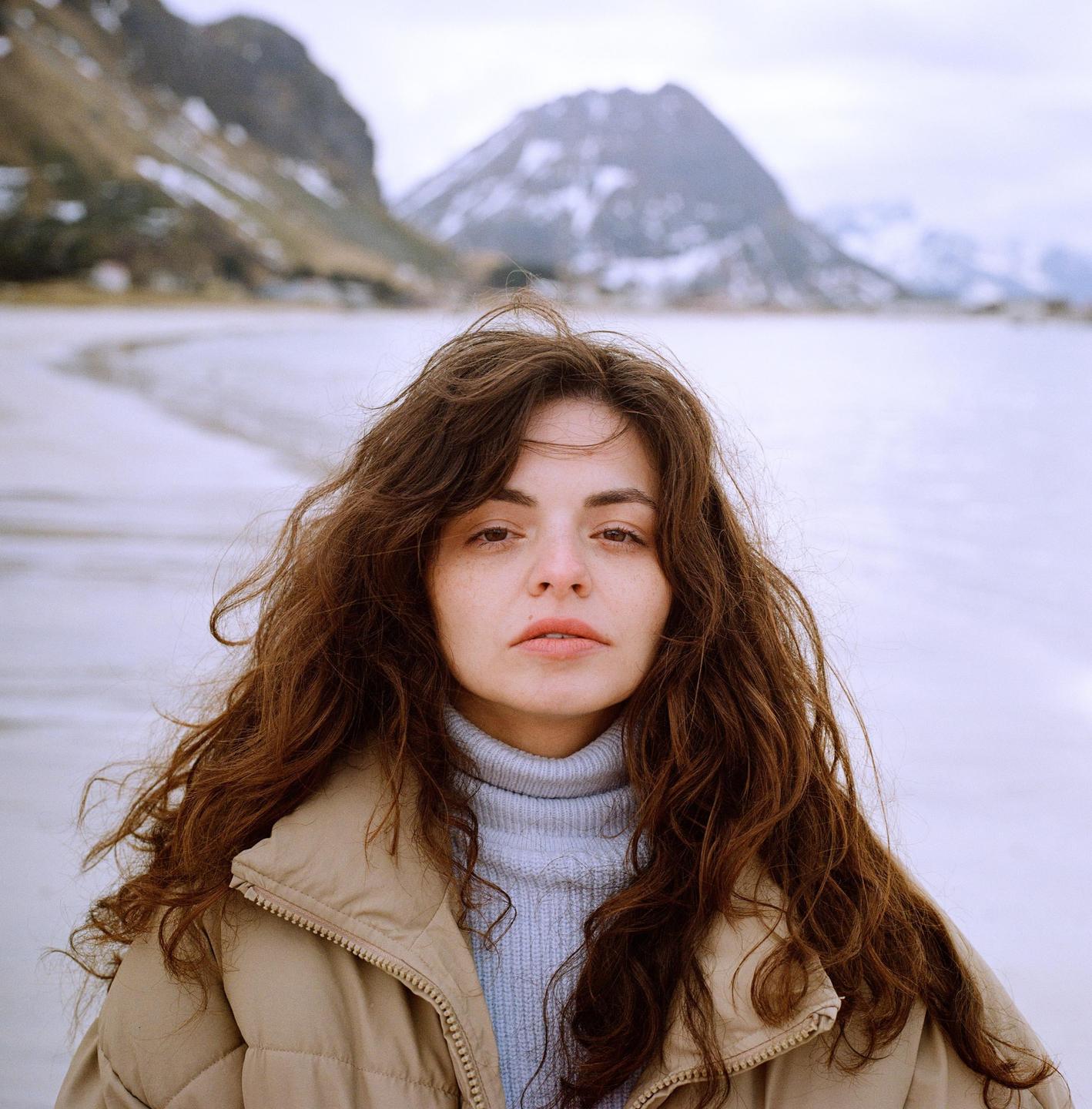 A woman with long, curly hair and a puffer jacket stands in a snowy landscape with mountains in the background.