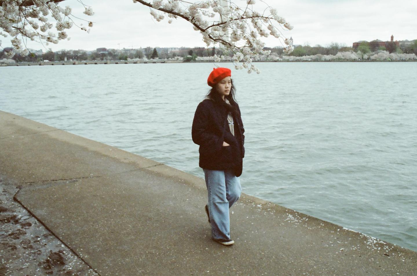 A woman in a red beret stands on a concrete path next to a body of water with cherry blossoms overhead.