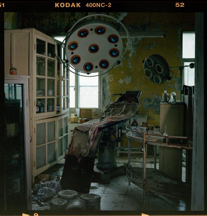 An abandoned operating room features a surgical table, medical lights, and cabinets with peeling yellow paint on the walls.