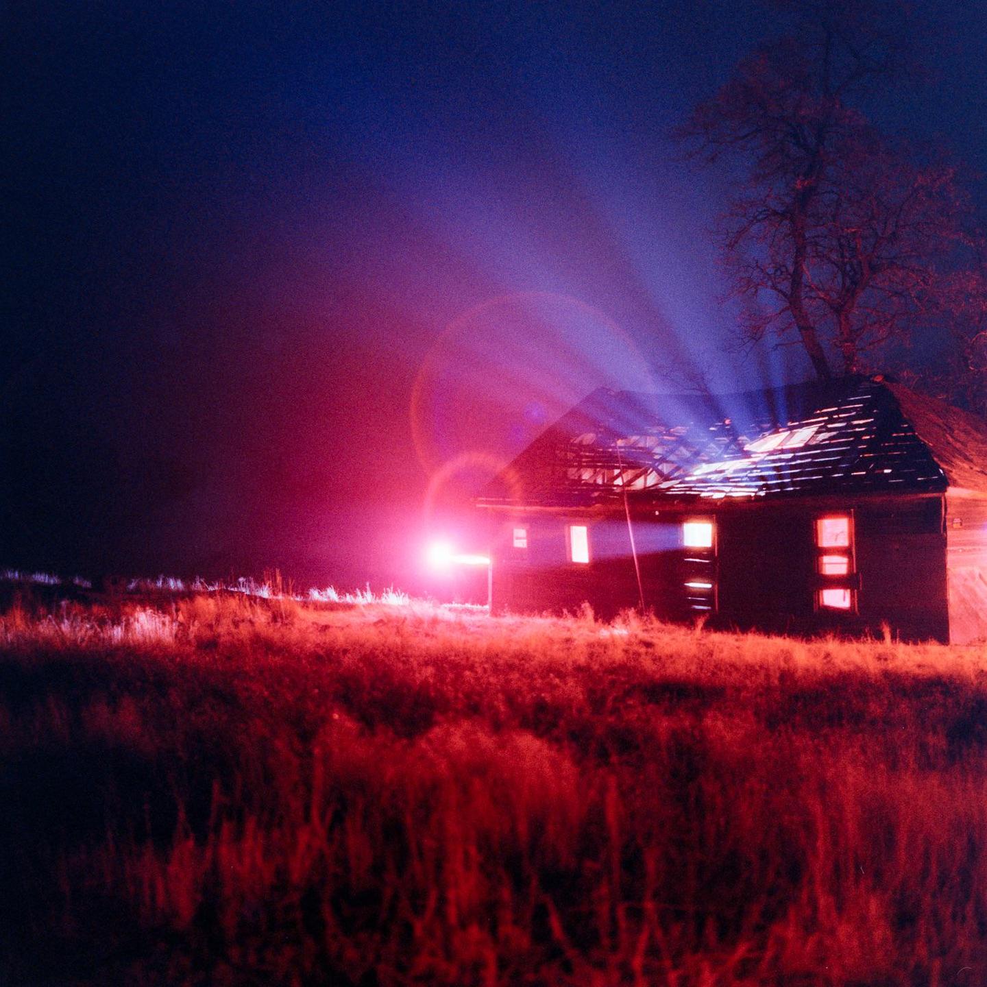 A house with a damaged roof stands in a field under a night sky with bright pink and blue light.