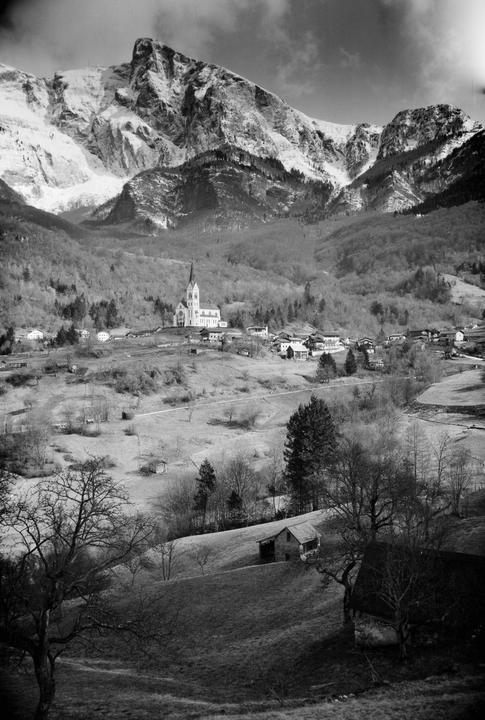 A village with a prominent church is nestled in a valley surrounded by snow-capped mountains and forested hills.