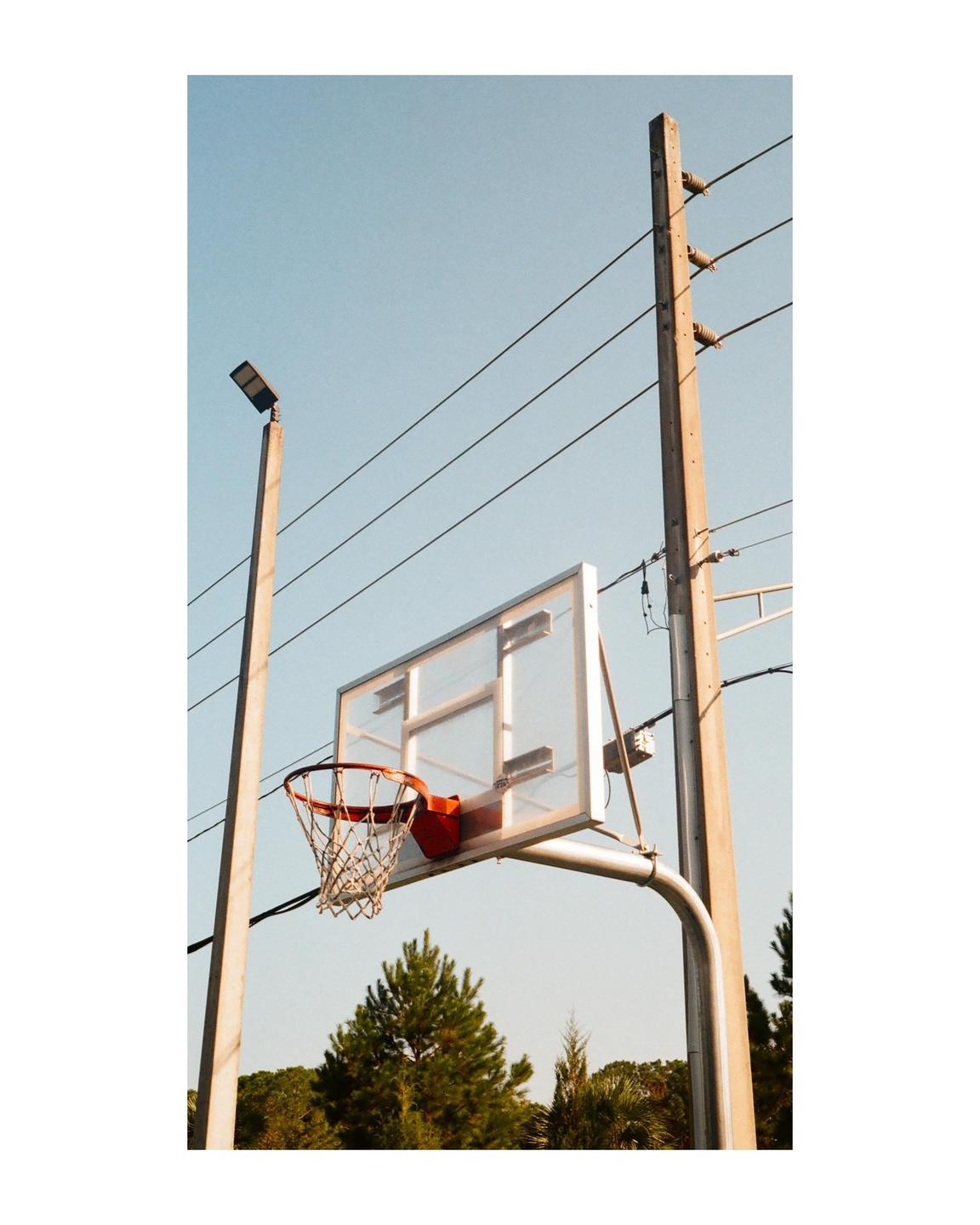 A basketball hoop with a net is visible against a clear sky, with utility poles and trees in the background.