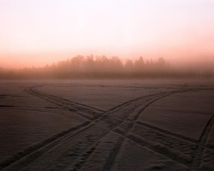 Tire tracks cross on a snow-covered field with a forest in the distance under a hazy, pink sky.