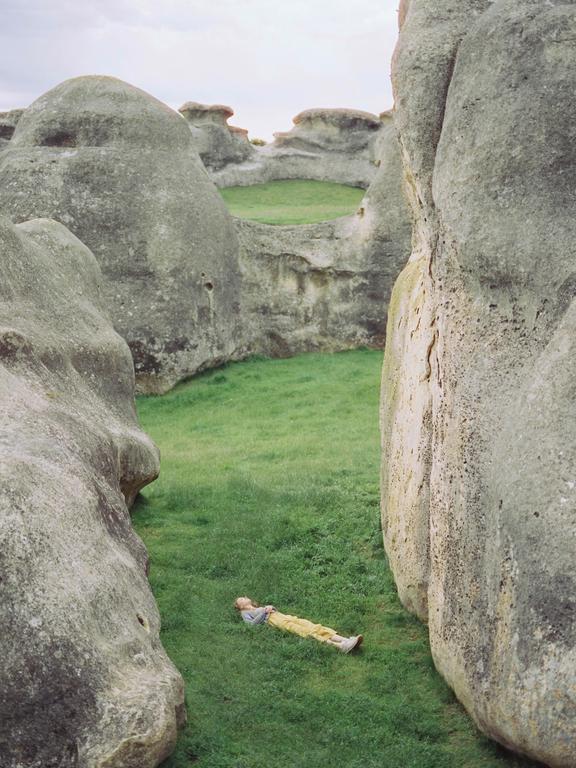 A person in yellow pants lies on green grass surrounded by large rock formations.